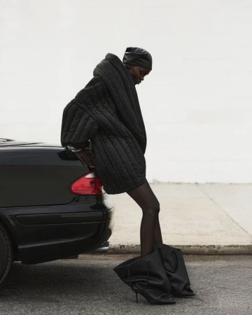 A slender woman wearing a black quilted jacket and matching headdress, leans on a black car parked on an outdoor city street.