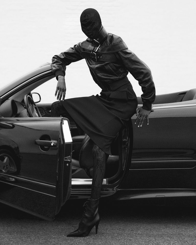 Black and white fashion portrait of an adult woman wearing a balaclava and leather jacket leaning against a convertible car.