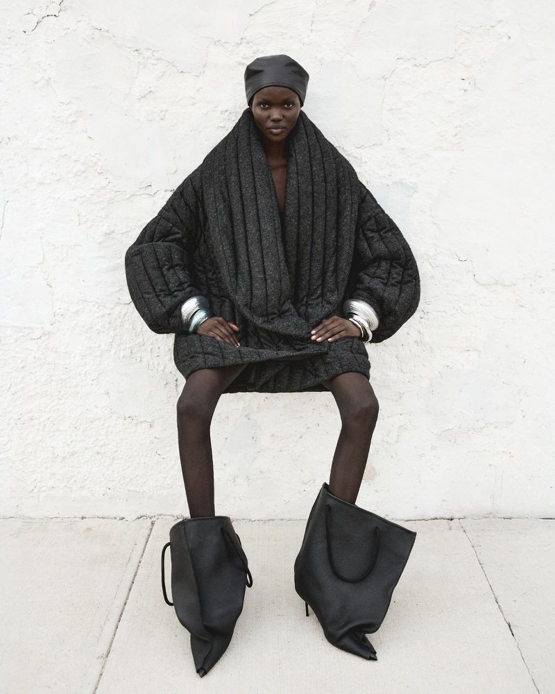 A fashion-forward individual with a dark complexion wearing an oversized black quilted coat, tights, and statement stiletto heels in an outdoor shot.