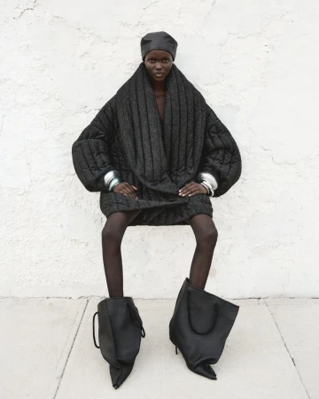A fashion-forward individual with a dark complexion wearing an oversized black quilted coat, tights, and statement stiletto heels in an outdoor shot.