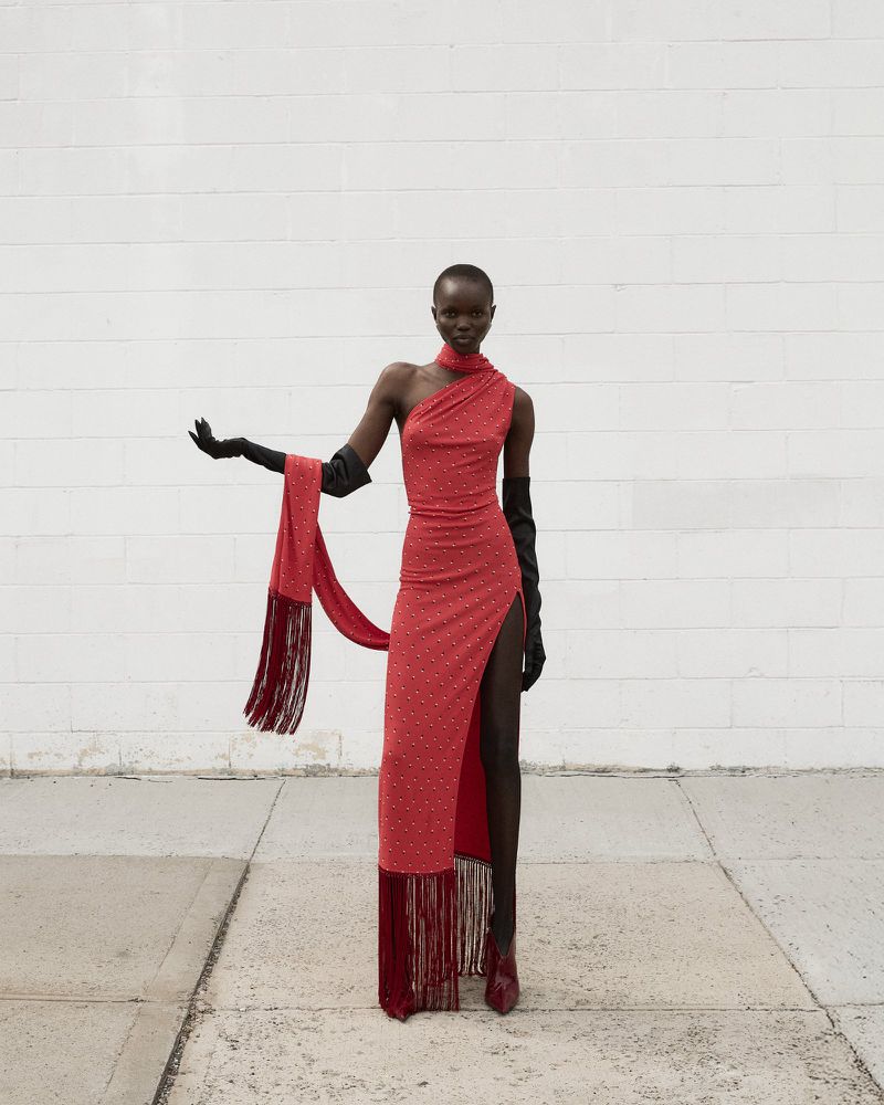 An adult female model poses in a one-shoulder red gown with a thigh-high split, wearing gloves in front of a white brick wall.