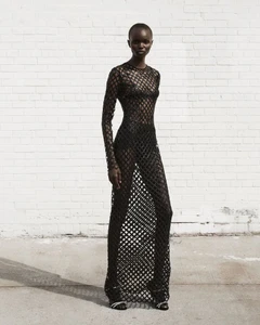 Model Adau Aguer poses in a sheer black open-mesh lattice gown against a painted white brick wall on concrete.