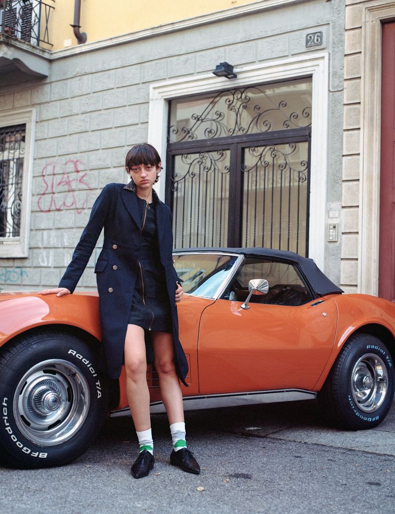A woman in a black coat poses next to a vintage orange convertible car outside a building on a sunny day in a fashion editorial.