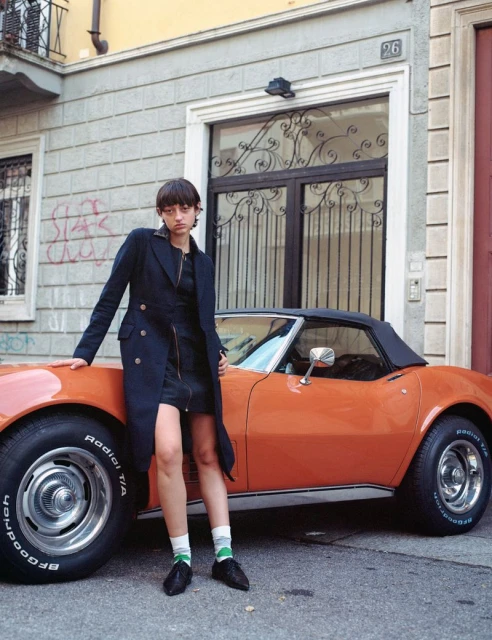 A woman in a black coat poses next to a vintage orange convertible car outside a building on a sunny day in a fashion editorial.