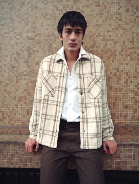 Asian man in a beige plaid jacket and brown pants stands against a mosaic tile wall, looking directly at the camera in a medium shot.