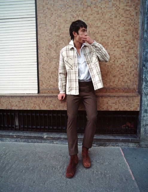 A man in a plaid jacket and brown pants leans against a tiled wall outdoors, giving a contemplative look, wearing suede boots.