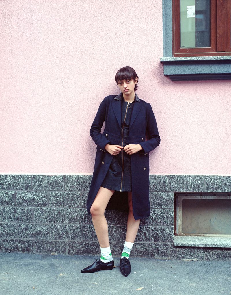 A full shot of a slender, fair-skinned person in a navy coat, black skirt, and patterned socks, posing against a pink and gray wall.