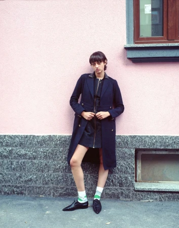 A full shot of a slender, fair-skinned person in a navy coat, black skirt, and patterned socks, posing against a pink and gray wall.