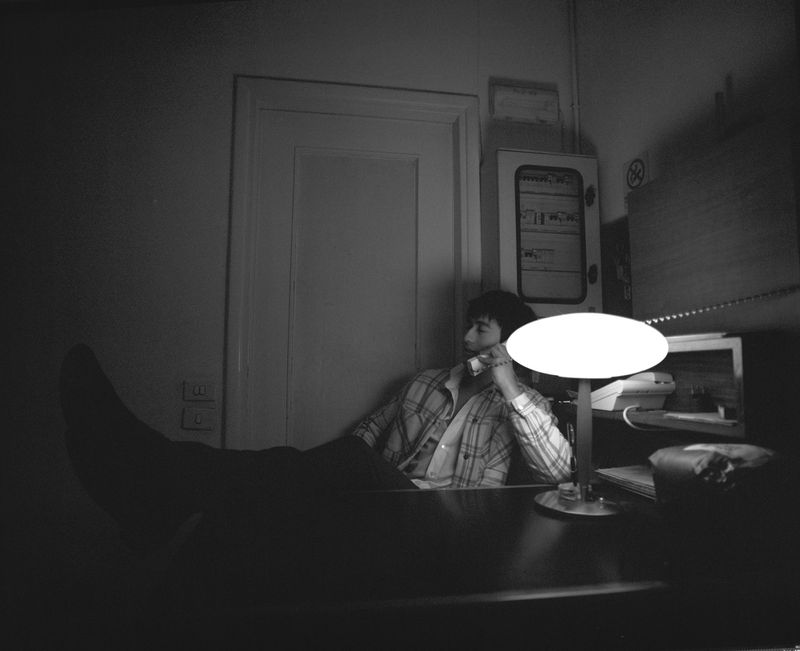 A man in a plaid shirt is sitting at a desk, talking on the phone under the light in an indoor setting in black and white.