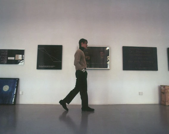 Man wearing a beige knit sweater and dark trousers walking in profile past framed artworks in an art gallery.