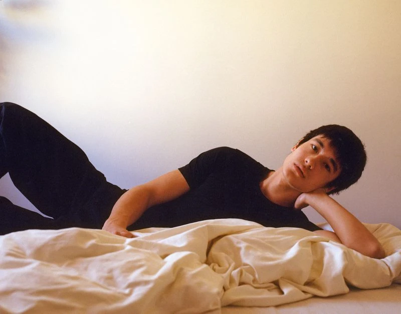 Young adult male poses on bed, laying down with arm resting near head. Soft daylight indoors with white walls, simple setting.