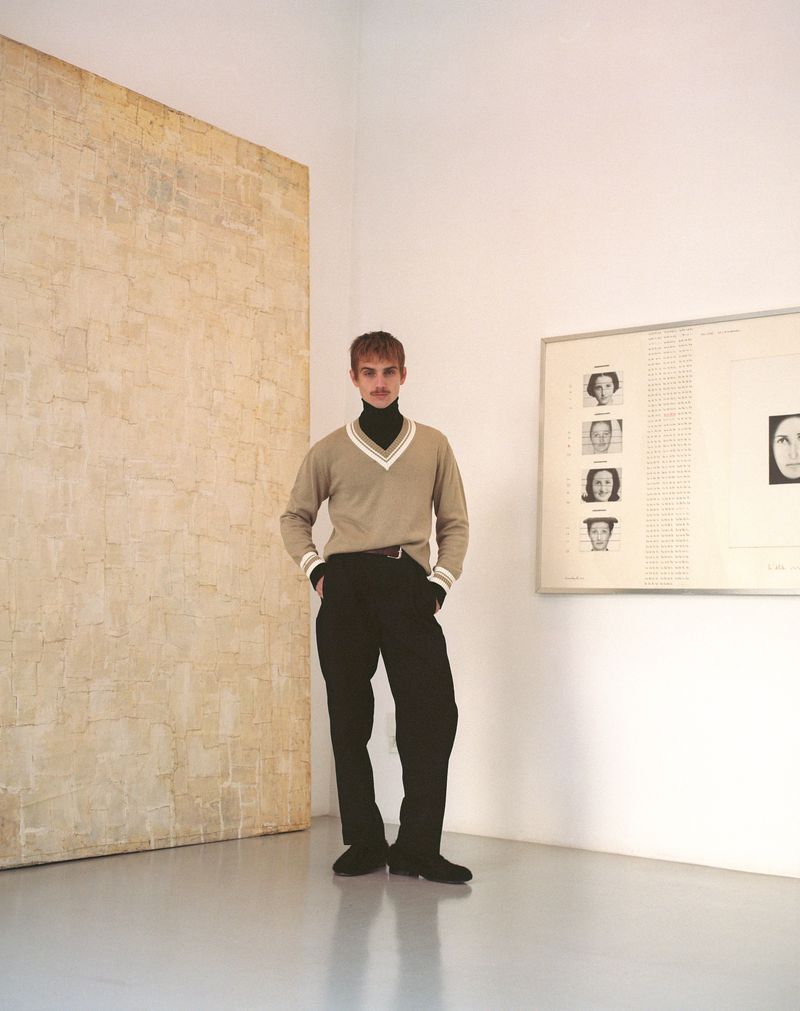 A young adult male stands in a sparsely decorated, light-filled studio, wearing a V-neck sweater and dark trousers, with artworks on the walls.