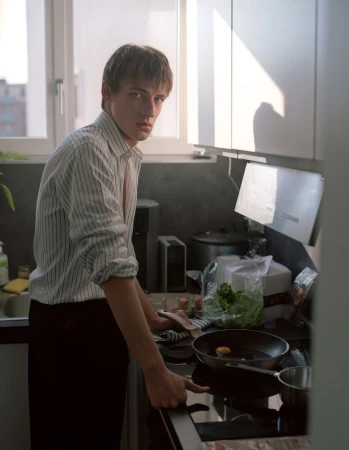 Young adult male stands indoors cooking at a kitchen stove; he wears a striped shirt and dark pants, with a sunny window behind.