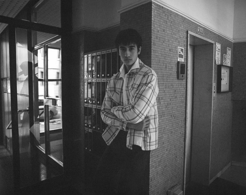 Black and white portrait of a young man with crossed arms, standing indoors near mailboxes and an elevator in a tiled corridor.