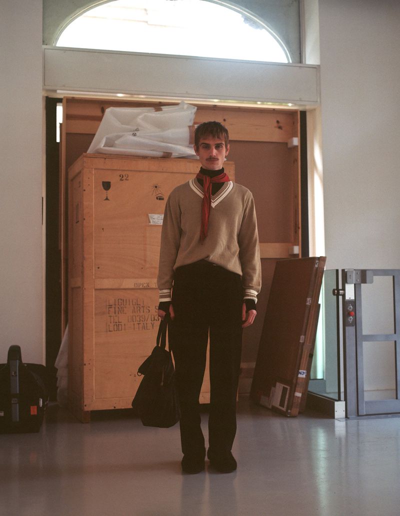 A casually dressed young adult male stands in front of a large wooden crate wearing a beige sweater, black pants, and a red scarf.