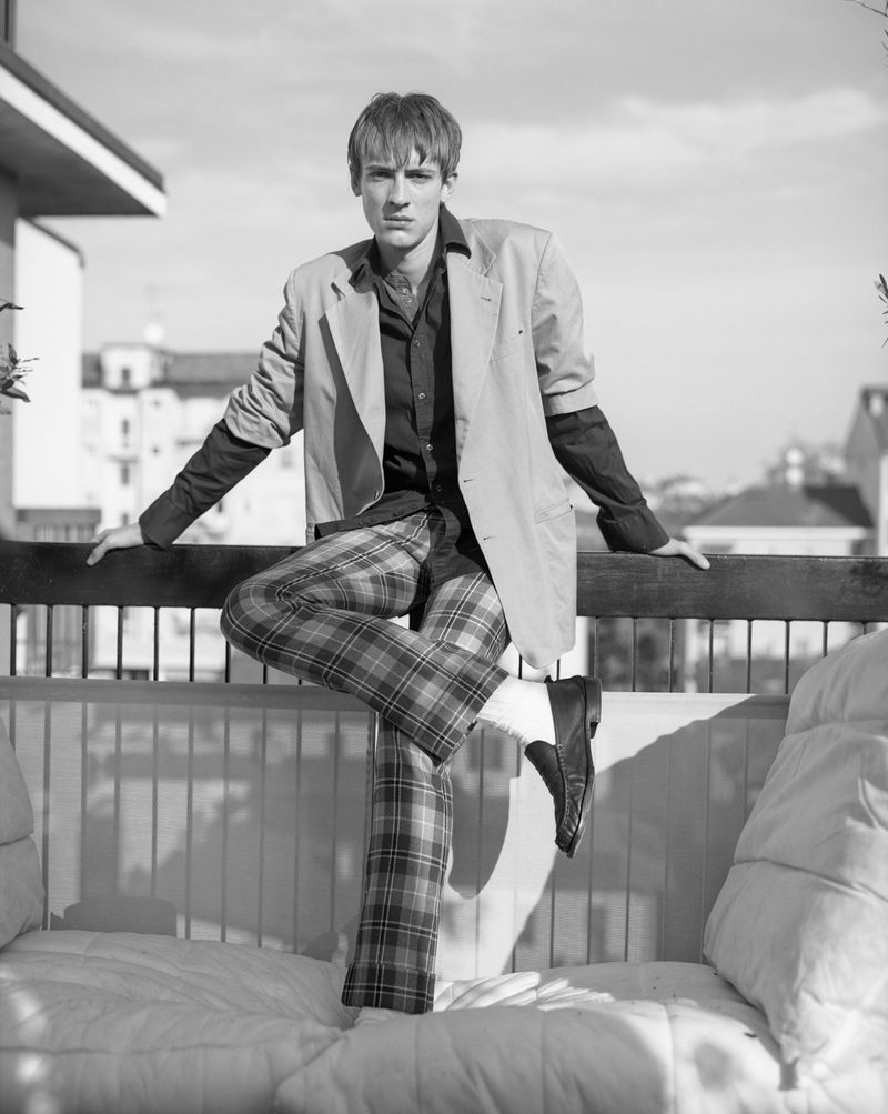 Black and white portrait of a young adult male in jacket and plaid pants posing outdoors on a cushioned bench with a city background.