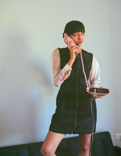A young woman with short dark hair stands indoors, holding a pink retro telephone to her ear and wearing a short, dark denim dress.