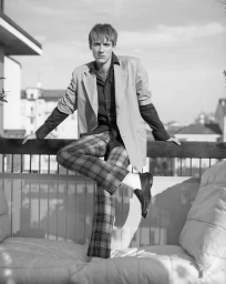 Black and white portrait of a man in an oversized blazer and plaid trousers leaning on a rooftop terrace railing.