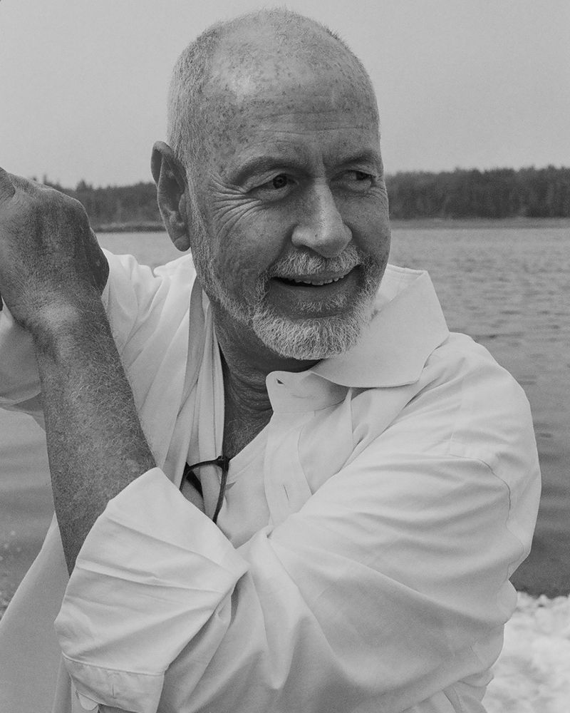 Black and white portrait of an elderly man with a beard wearing a white collared shirt against a background of water and trees.