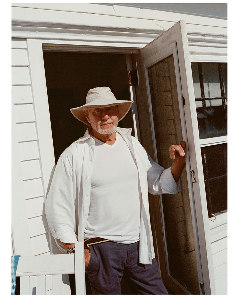 An older man with a beard and hat stands in an open doorway, wearing a white shirt and blue pants, on a sunny day, looking at camera.