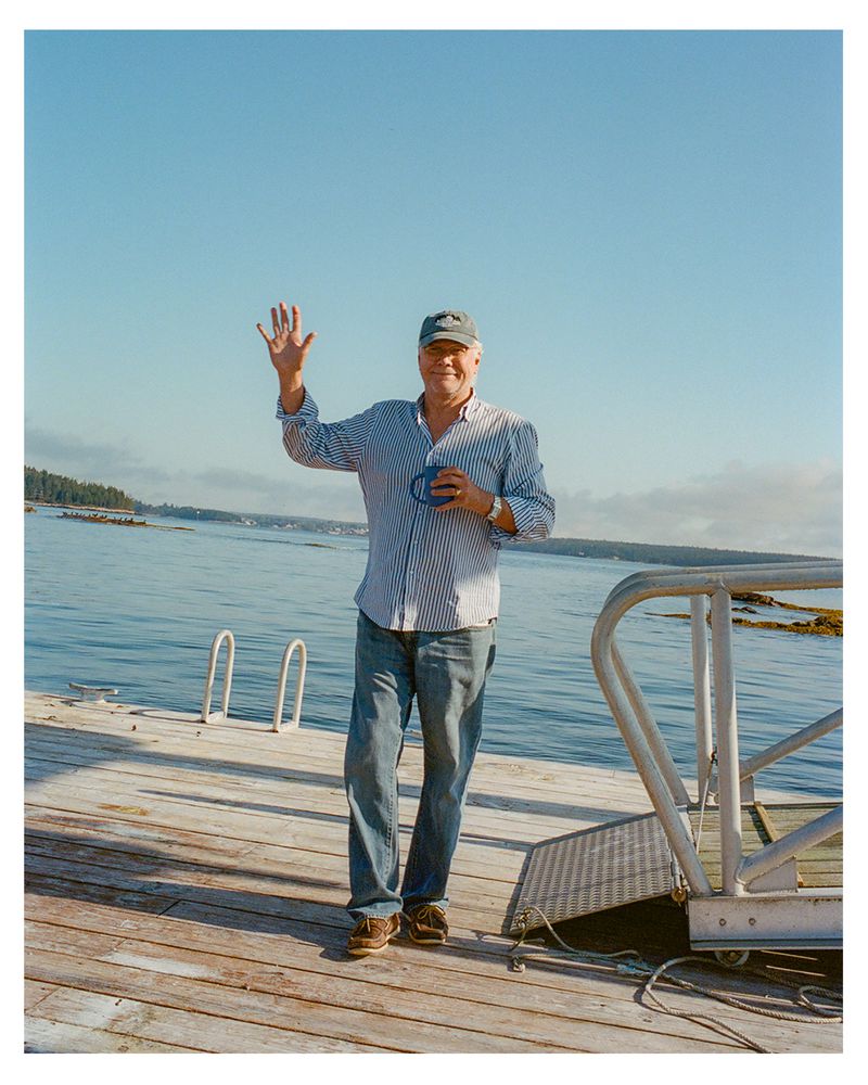 Mature adult man waving on a wooden pier near the ocean, wearing a striped shirt, jeans, and cap, holding a mug on a sunny day.