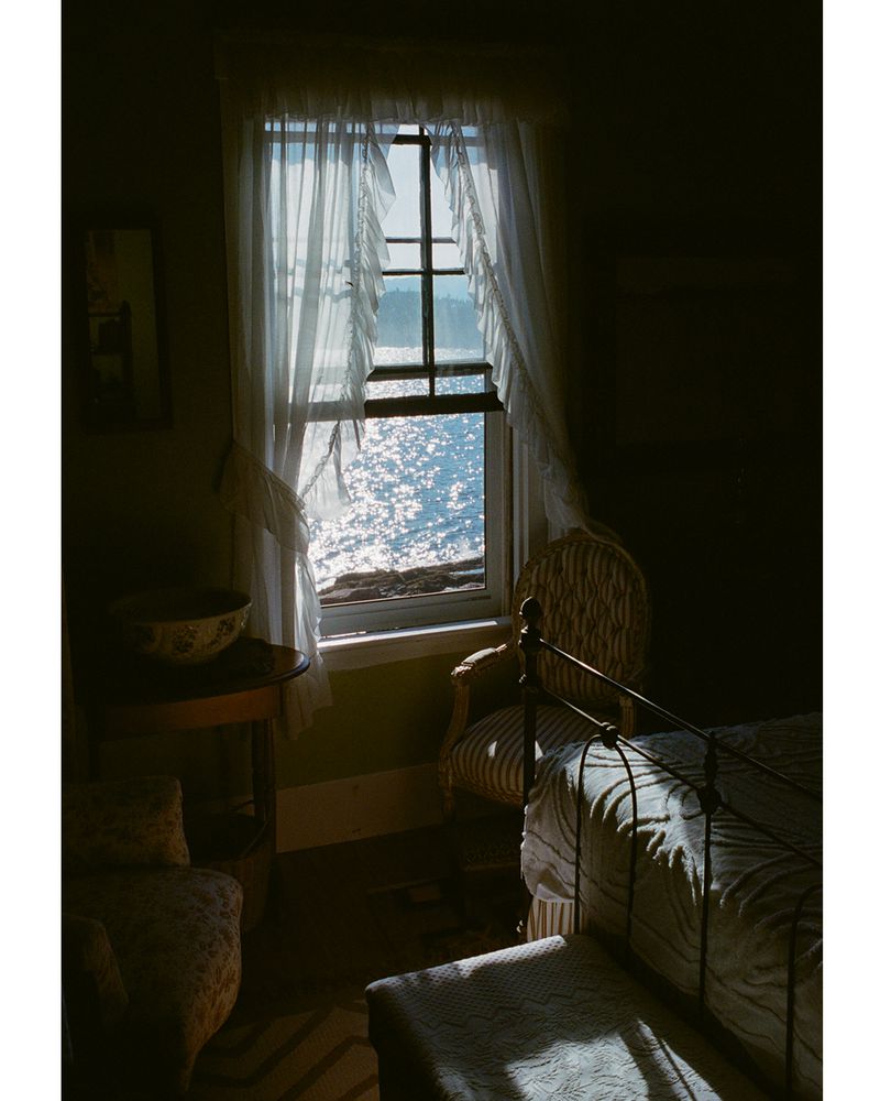 Sunlight streams through window with sheer curtains into a vintage bedroom with bed, chairs, and glimpse of the ocean.