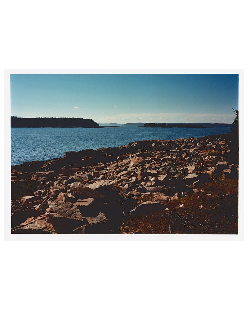 Coastal landscape featuring a rocky shoreline with large stones and calm blue water under a clear sky, showcasing natural beauty.