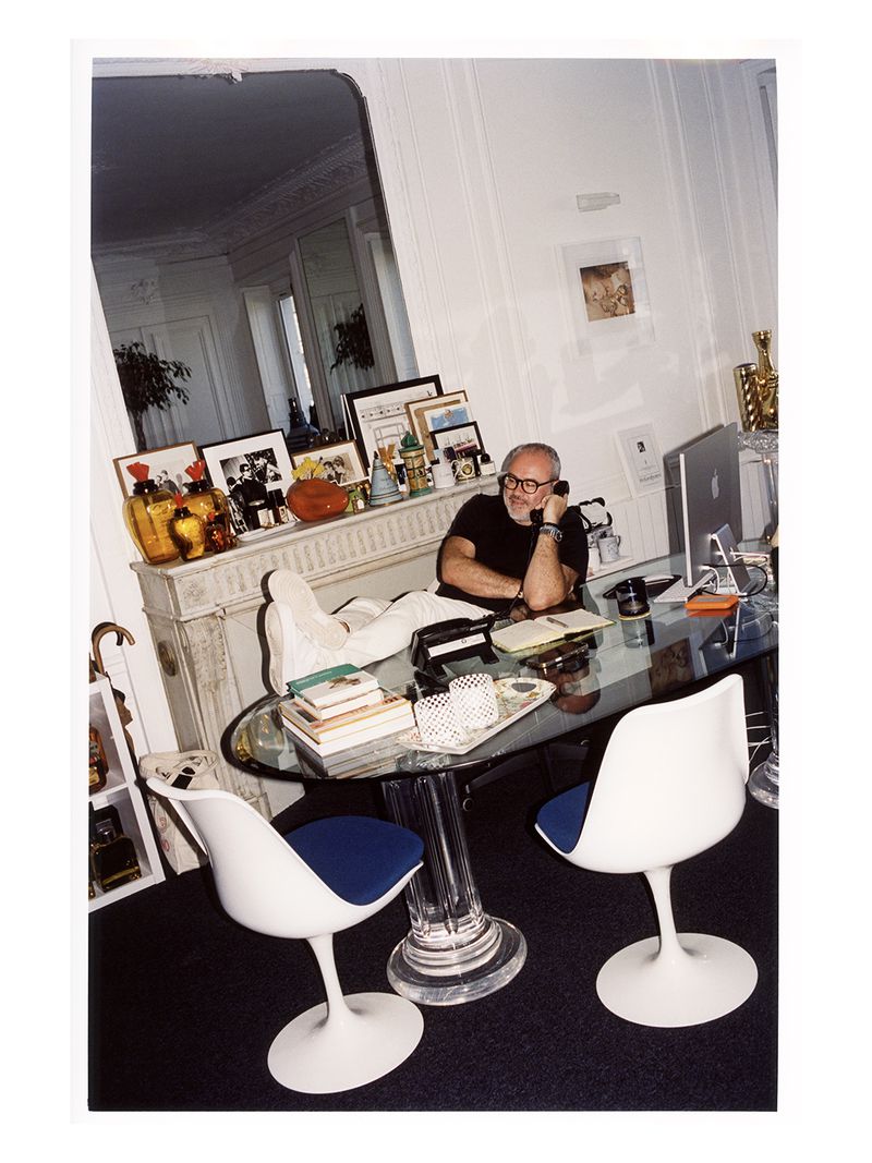 Adult man in an office setting, wearing glasses and talking on the phone while seated at a glass desk with papers and books.