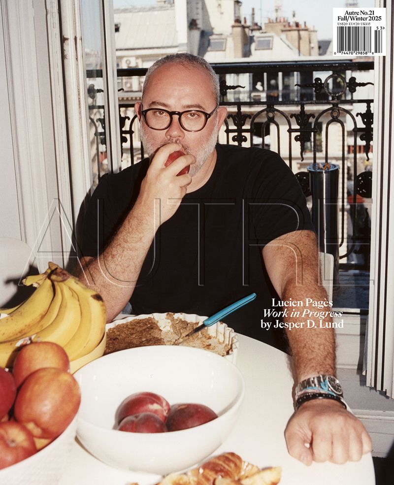 Adult man with glasses eating an apple at a white table in front of window with cityscape view; bananas, peaches, croissants visible.