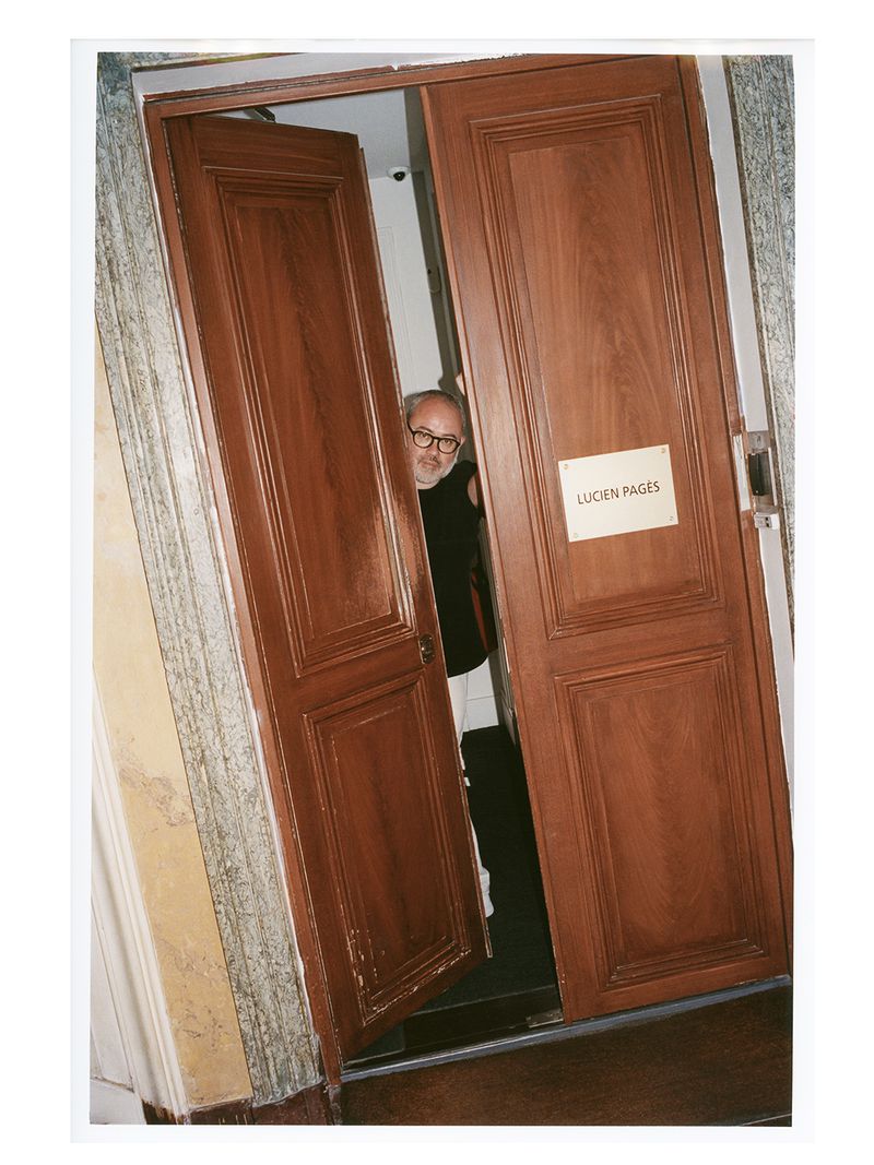 An adult male with glasses peers through a wooden doorway with a sign marked Lucien Pages, inside a marble-walled building.