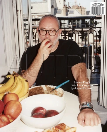 Man wearing glasses and black t-shirt eating an apple at a table with fruit and a pastry in front of a window.