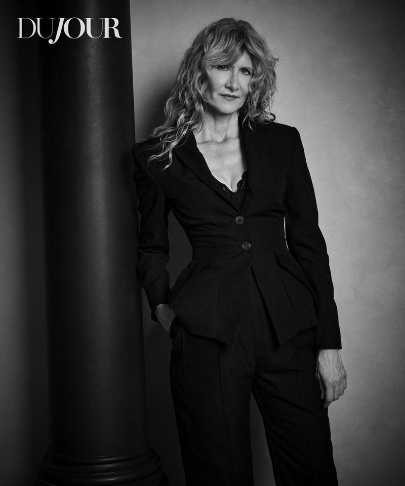 Black and white portrait of an adult woman with wavy hair wearing a suit, leaning against a column in a photo studio.