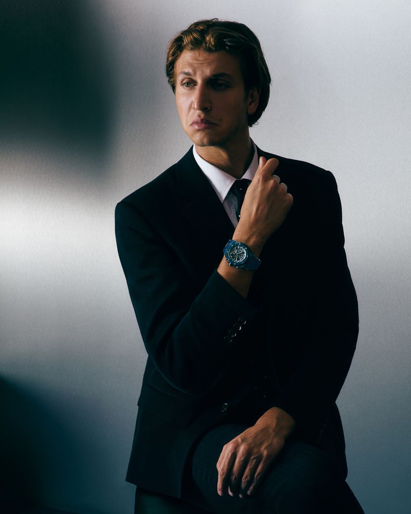 Adult male in a black suit and white shirt posing indoors in front of a light gray wall while wearing a blue wristwatch.
