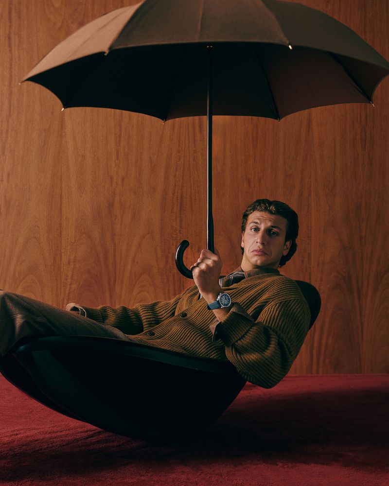 Stylish man sitting in a chair under an umbrella, wearing a cardigan sweater, wrist watch, and dark pants against wood panel background.