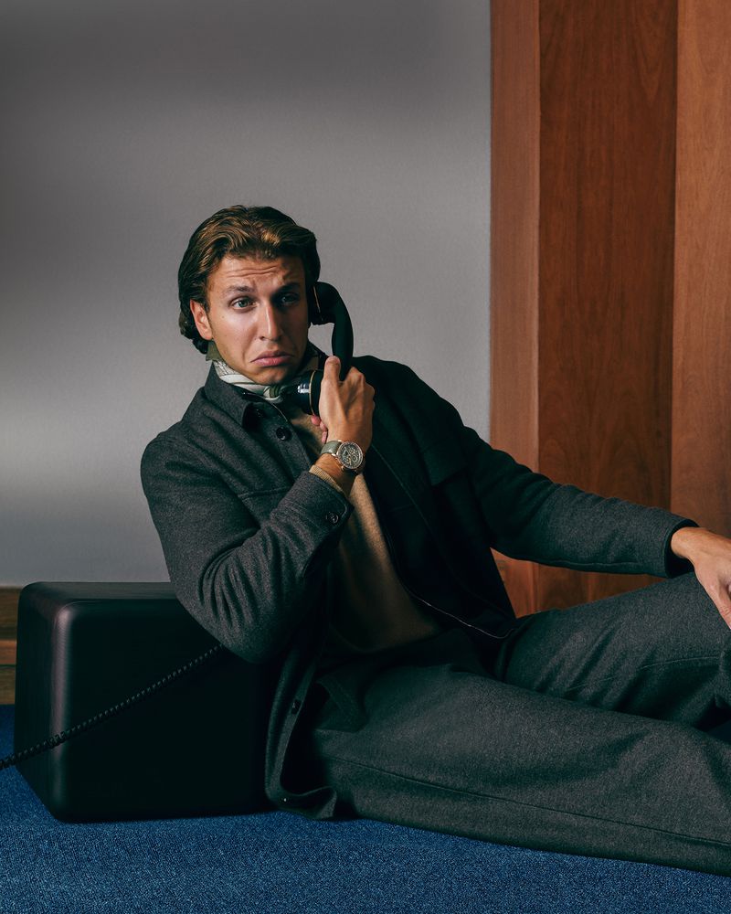 Man in a gray suit sitting on a black block while using a vintage telephone in an indoor setting with wood paneling.