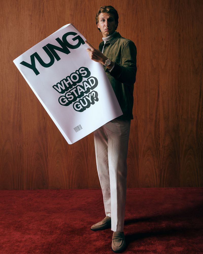 Adult male in a green jacket and cream pants stands holding a poster with the text YUNG, wearing a watch, in a wood-paneled room with red carpet.