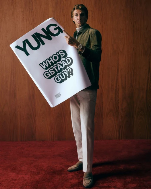 Adult male in a green jacket and cream pants stands holding a poster with the text YUNG, wearing a watch, in a wood-paneled room with red carpet.
