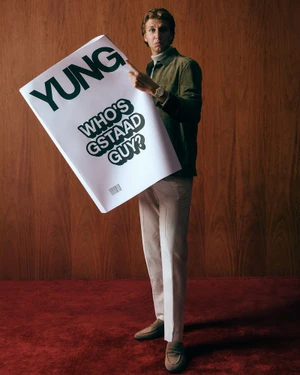 Man holding a large magazine pointing at the cover against wood paneling and red carpet