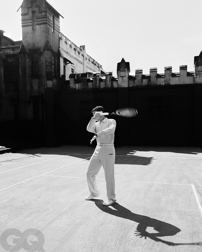 A man in athletic wear is poised to hit a tennis ball with a racquet on a tennis court. The background is a tall brick building.