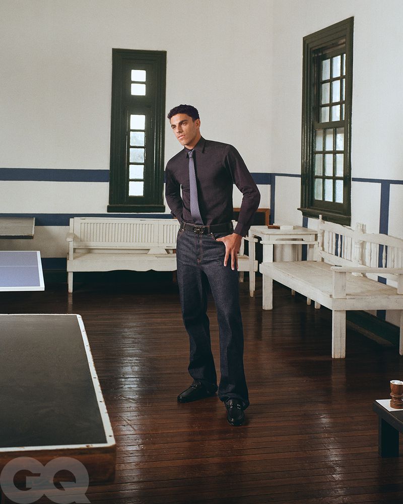 Portrait of a stylish adult male in a black shirt and dark denim jeans posing indoors with ping pong table and white benches.