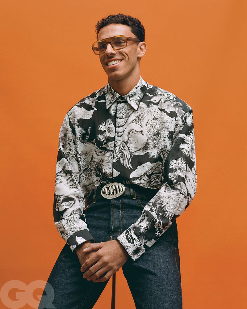 Portrait of a smiling adult male in an animal-print shirt, wearing glasses and a Moschino belt, against an orange background.