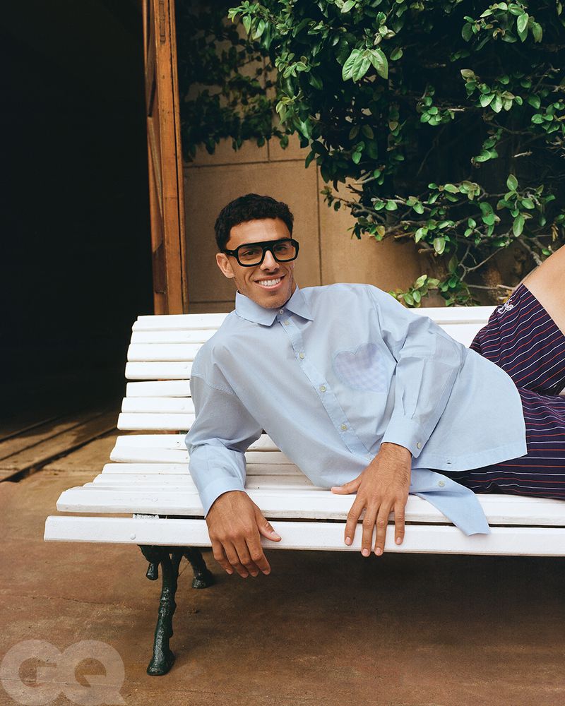 Smiling man in glasses and shirt lounges on a bench outside near greenery. Light blue button-down, striped shorts, relaxed pose.