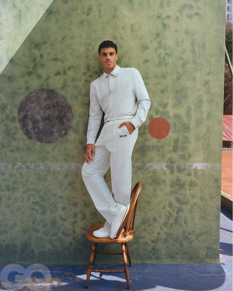 An adult male model in a white pantsuit standing on a wooden chair in front of a green wall and outdoor tennis court for a fashion shoot.