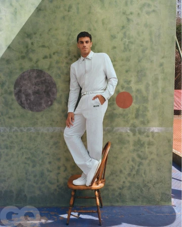 An adult male model in a white pantsuit standing on a wooden chair in front of a green wall and outdoor tennis court for a fashion shoot.
