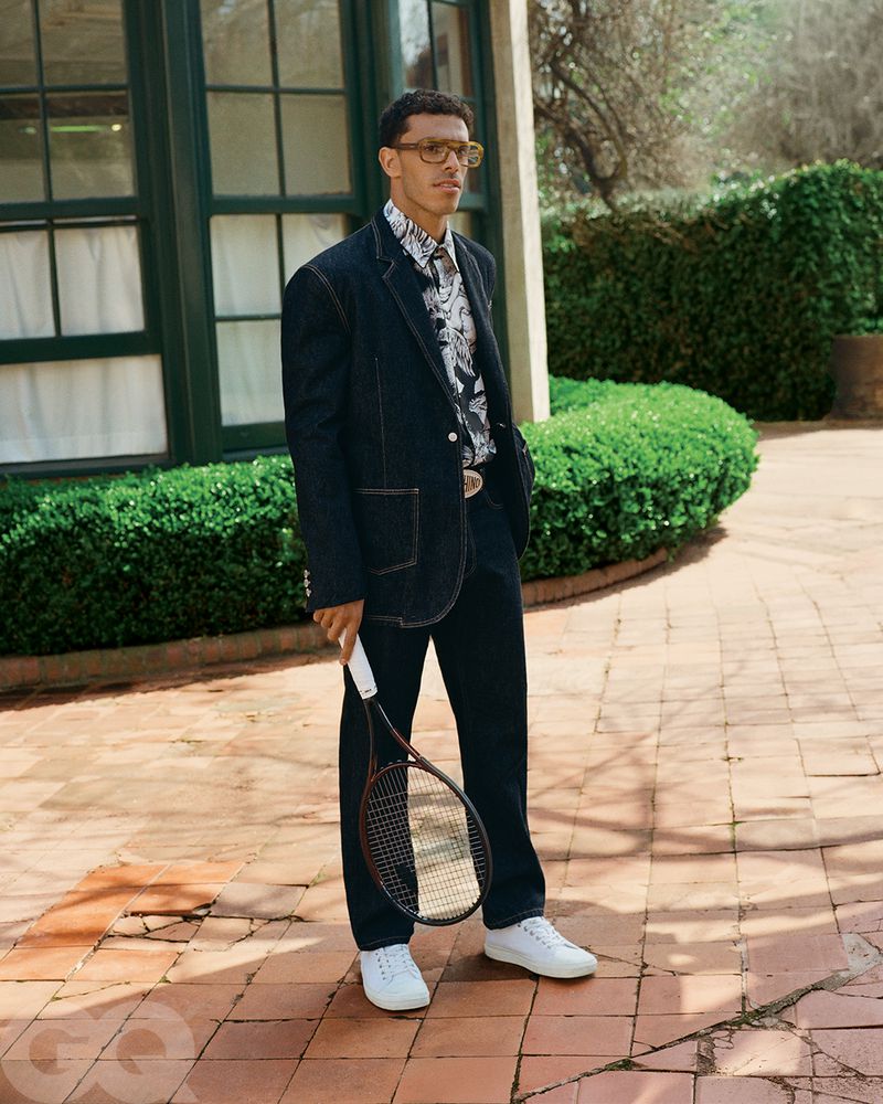 Man poses in a denim suit, patterned shirt, and white sneakers, holding a tennis racket against an outdoor backdrop of greenery and brick.