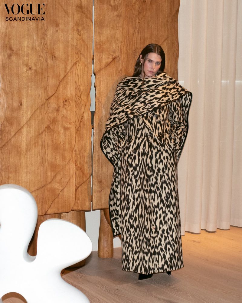 Fashion shot of a woman in a patterned coat standing in an indoor environment with light wood panelling and a white flower sculpture.
