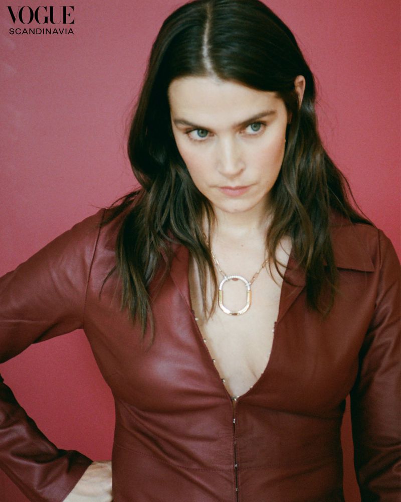 Brunette woman in a burgundy leather top and gold necklace posing in front of a pink backdrop for a fashion editorial portrait.