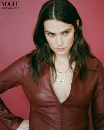 Brunette woman in a burgundy leather top and gold necklace posing in front of a pink backdrop for a fashion editorial portrait.