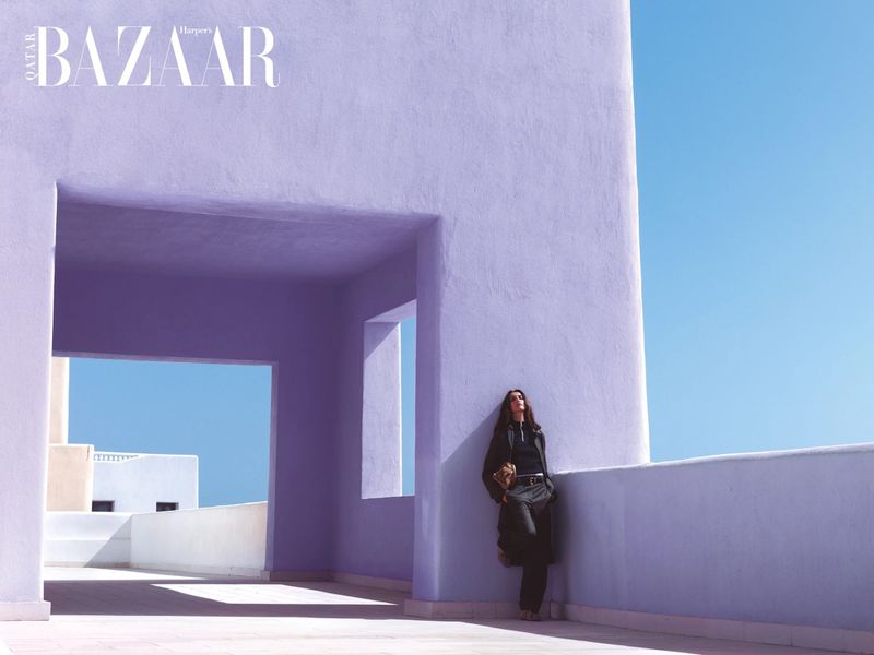 Fashion shot of a woman in dark clothing leaning against a lavender building under a bright blue sky, with magazine logo in the upper left.