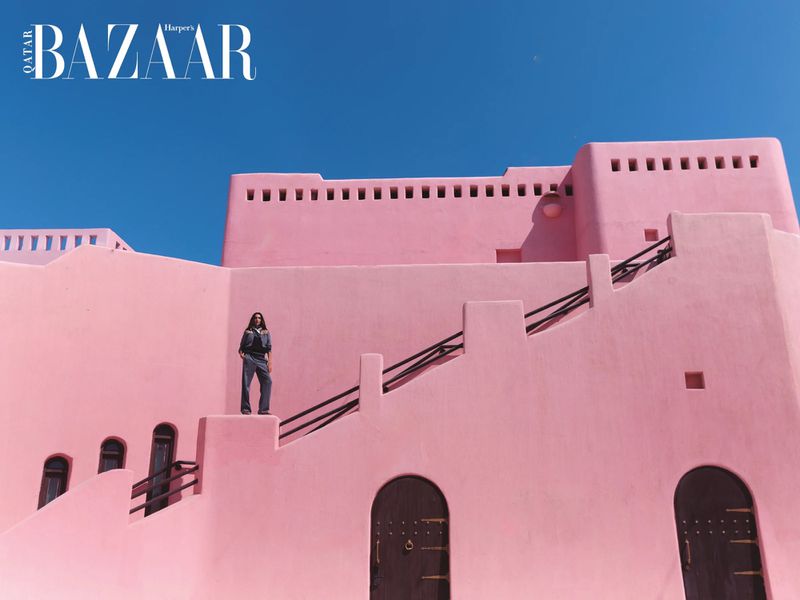 Fashion editorial shot of a model posing on the steps of a pink, historic building under a clear blue sky for Qatar Bazaar.